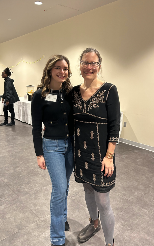 “Two individuals stand side by side in an indoor event space. According to the user, the person on the left is Anna Russo (PC Class of 2021, WGS minor, Accounting major), and the person on the right is Dr. Brooks. The individual on the left is wearing a dark buttoned jacket and blue jeans. The individual on the right is wearing a black dress with decorative embroidery, light-colored tights, and dark shoes. A table with refreshments is visible in the background, along with a few other people in the room.”
