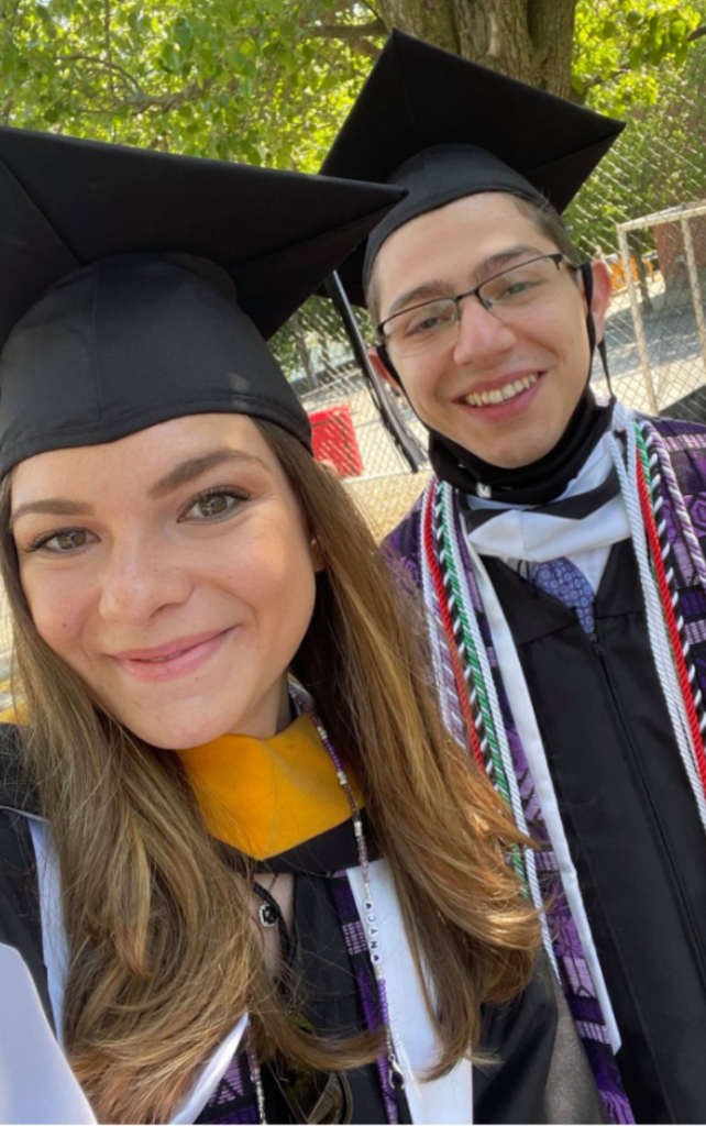 “Two individuals wearing graduation caps and gowns stand close together outdoors, with a tree and a chain‑link fence in the background. According to the user, one individual is PC WGS Minor and PSC Major alum Chris Iadeluca (Class of 2021). Both individuals are dressed in academic regalia with stoles and cords visible. The photo is taken as a close-up selfie.”