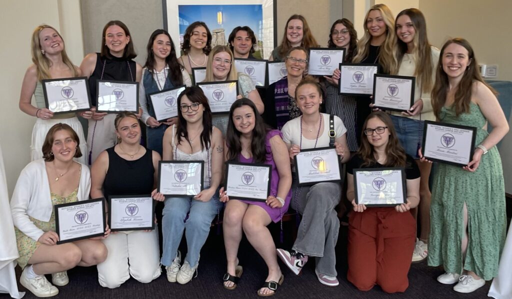 A group of 19 individuals poses indoors for a formal photo, holding certificates featuring a purple and white emblem. They are arranged in three rows, with some seated in front and others standing behind. The setting suggests an award or graduation ceremony, with a framed picture and two wall-mounted lights visible in the background.